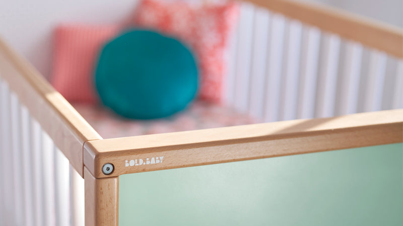 Bold Baby logo on corner of cot with red and green cushions in the background