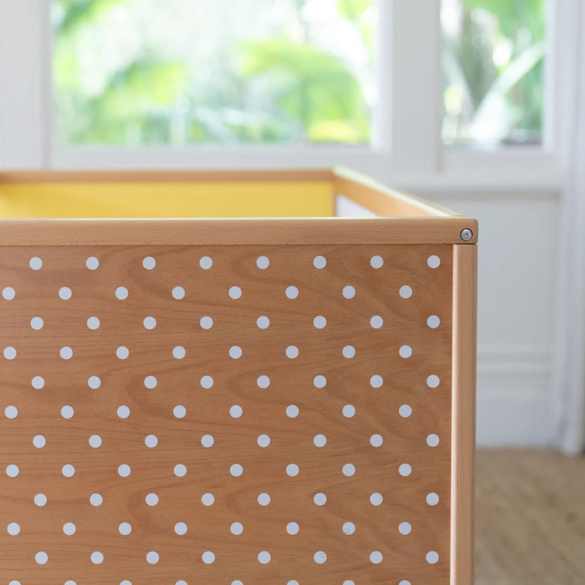 Close-up of a wooden storage bin or crib with a natural wood finish and evenly spaced white polka dots on the front panel, set against a softly blurred indoor background with a window.
